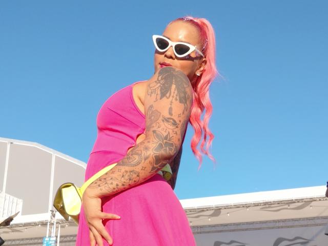 Chili, a busty, tattooed milf with pink hair and white sunglasses, stands in a bright pink strapless dress, touching her hip.
