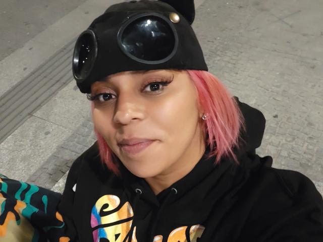 Chili, milf solo, avec des cheveux roses, portant un bonnet noir et des lunettes sur sa tête, sweat à capuche noir, fond de trottoir de ville.