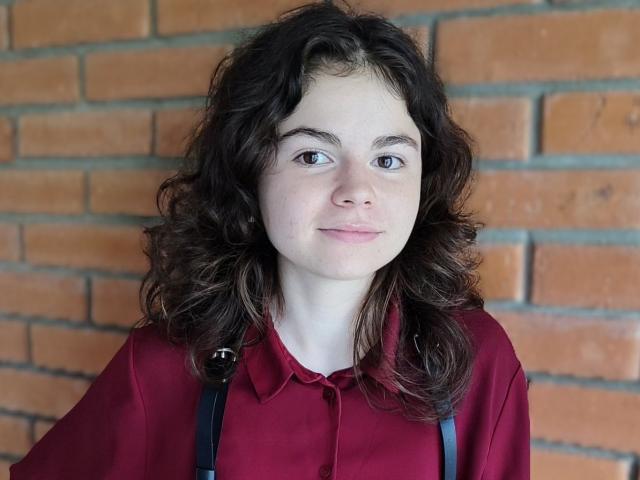 TinaRedly has curly brown hair, light skin, and blue eyes, wearing a red shirt with black suspenders, smiling slightly.