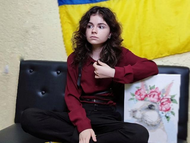 Tina, mit lockigem, braunem Haar, trägt einen roten Pullover und eine schwarze Hose und sitzt auf einem schwarzen, gepolsterten Sofa. Sie hält ein Porträt eines Hundes mit rosa Rosen..