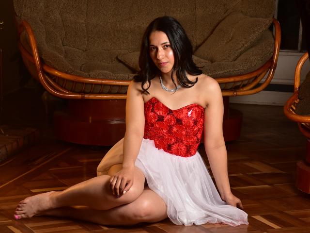 Anne Stone, con i capelli neri, indossa un abito rosso in pizzo e una gonna bianca, seduta su un pavimento di legno, con le gambe incrociate, sorridente, a piedi nudi, con una collana, una sedia marrone sullo sfondo..
