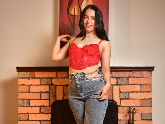 AnneStone with dark hair, red lace top, showing small tits, and blue jeans, smiling, hand on hip, standing in front of a brick fireplace, necklace, sexy.