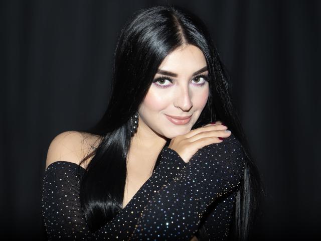 Ariana Cortes, com cabelos longos e pretos, vestindo uma blusa preta com detalhes em preto e branco, com um ombro de fora, sorrindo, com as mãos perto do rosto, em um fundo escuro, parecendo adorável com seus olhos rosa..