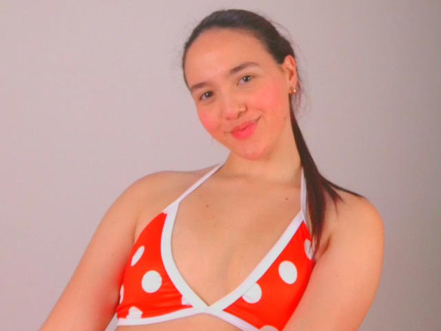 Milliee, usando um top de biquíni vermelho com bolinhas, que mostra seus seios, tem um leve sorriso, cabelo castanho preso em um rabo de cavalo e um tom de pele claro, contra um fundo simples..
