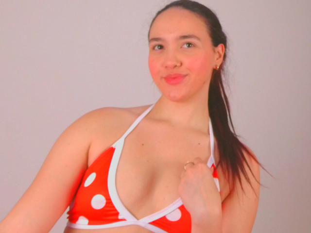 Milliee with red polka dot bikini top, showing her small tits, has a light skin tone, brown hair in a ponytail, and a slight smile, against a plain background.