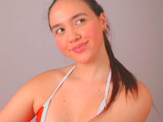 Milliee, con una coleta oscura, labios rosados, un piercing en la nariz, pequeños senos en un top de bikini rojo y blanco, una ligera sonrisa, piel clara, pequeñas pecas marrones, mejillas rosadas, aretes dorados, luz suave..