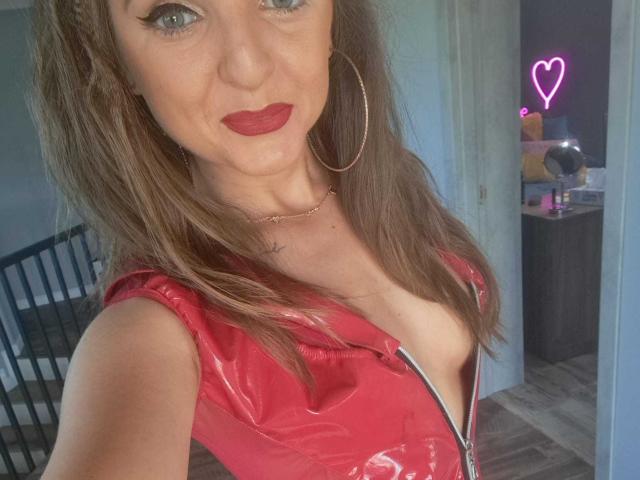 WildBlondy69 with red vinyl zipper top, showing cleavage, wearing hoop earrings and red lipstick, brown hair, smiling, in a well-lit room with a neon heart in the background.