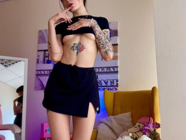 Tsukerberg con piccoli seni, top nero, gonna nera, tatuaggi, dito in bocca, in piedi, tatuaggio di una rosa visibile sul petto, sedia gialla sullo sfondo, riflesso nello specchio a sinistra..