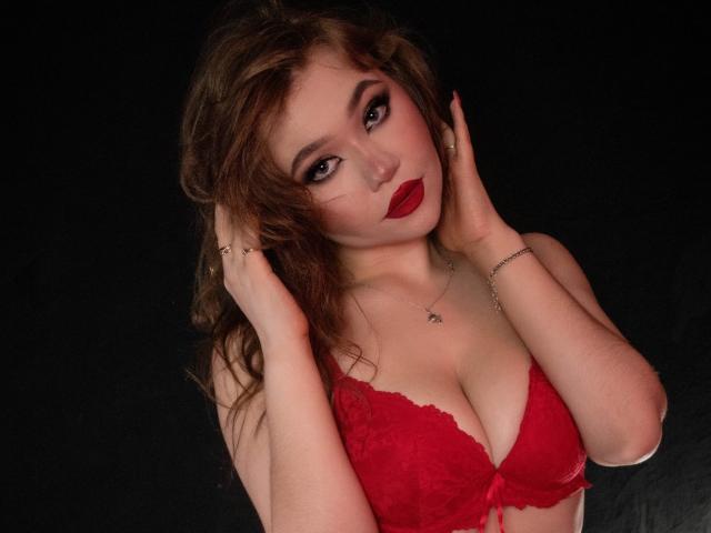 Stacy Yellig com um sutiã de renda vermelho, mostrando seus seios, mãos no cabelo castanho bagunçado, batom vermelho e fundo preto, olhando para cima com um leve sorriso..