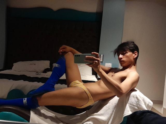PiesoLores, topless with yellow panties, blue knee socks, and short black hair, lounges on a bed, taking a mirror selfie, showing a slim, toned body and a small ass.
