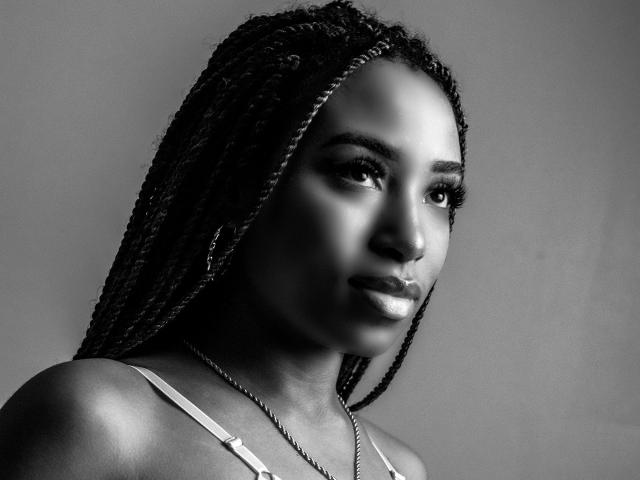 Photo en noir et blanc de SiaHotty avec de longs cheveux tressés, portant un haut à bretelles fines, regardant sur le côté avec un léger sourire, et portant un collier en chaîne..