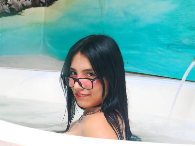 TiffanyEve with black hair, wearing glasses, in a bathtub, showing her tits, smiling, with water up to her shoulders, and a silver necklace.