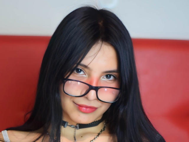 TiffanyEve tiene el pelo negro, ojos azules y usa gafas. Está sonriendo con lápiz labial rojo, lleva un collar negro y una blusa blanca, y se ve linda con un fondo rojo..