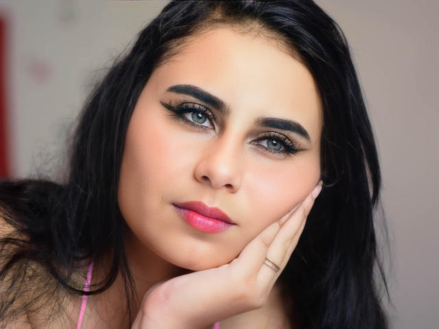 LeilaEvanns with black hair, blue eyes, pink lipstick, and dark eyeliner, resting her chin on her hand, showing her pink top, with a slight, sexy, confident smile.