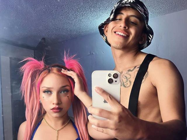 Pinkandkey69, avec ses couettes roses, son haut bleu et son maquillage, se tient à côté de Pinkandkey69, torse nu, portant un chapeau, des tatouages et des bretelles, et ils prennent un selfie avec un iPhone..