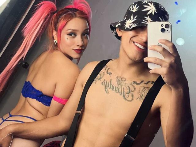Pinkandkey69 met roze vlechtjes, een blauwe kanten bh en een roze string, waarbij haar billen te zien zijn. Pinkandkey69 zonder shirt, met tatoeages, braces, een vissershoedje, lachend en een telefoon vasthoudend..
