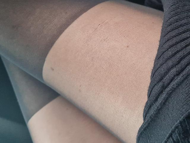 ApoloniaRed's legs in sheer black stockings, showing a small tear on the upper thigh. Her skin is light with some small dark spots. She's wearing a black dress.