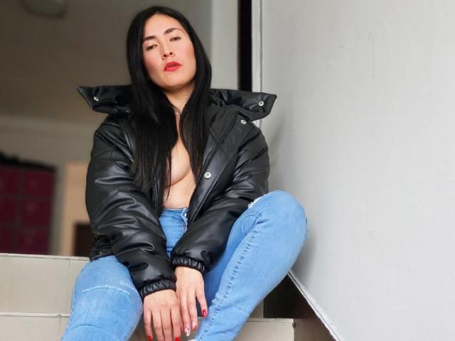 JainaRogers with long black hair, wearing a black puffy jacket, no top, exposing small tits, and blue jeans, sitting on stairs, looking slightly to the side.