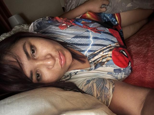 KarlaWild ligt op een bed, gekleed in een Spider-Man shirt, met bruine ogen, een medium huidskleur, donker haar, lichtjes open lippen, en kijkt naar de camera, met een rode deken..