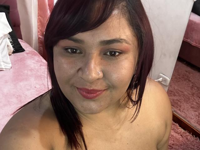 KarlaWild with medium brown skin, dark red hair, subtle makeup, pink lipstick, and a small nose ring, smiling at the camera, topless, showing her shoulders and a slight double chin.
