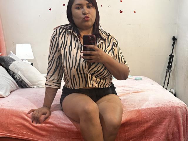 KarlaWild, con i capelli castani, una camicia a righe e pantaloncini neri, seduta su un letto rosa, con un'espressione accigliata, tiene in mano un telefono, mostrando le cosce e la biancheria intima nera..