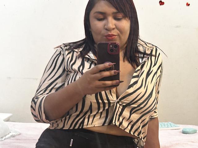 KarlaWild mit dunkelbraunen Haaren, einem gestreiften Hemd und rotem Lippenstift, hält ein Telefon, zeigt ihre Brüste, sitzt auf einem Bett, macht ein Selfie, trägt schwarze Hosen, leicht schmollend..
