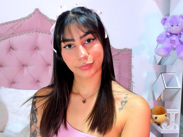 JessicaSoler has black hair, pink eyes, and a cute smile. She's wearing a pink top, showing tattoos on her shoulders. Her small tits are visible. Pink headband. Bedroom.