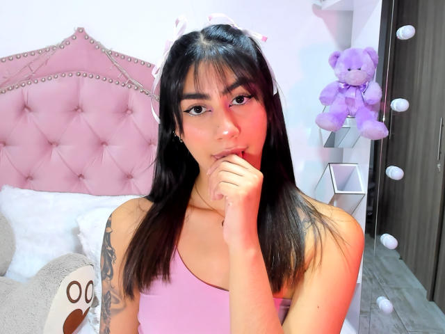 JessicaSoler with black hair, pink tank top, finger in mouth, tattoo on left arm, purple stuffed bear, pink tufted headboard, white wall, and mirror with round lights.
