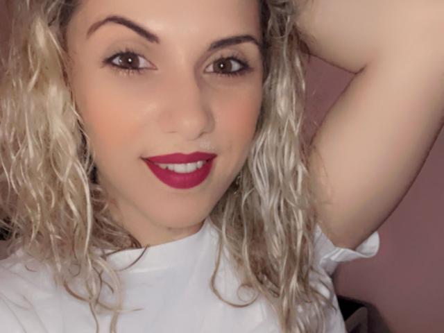 LucilleXSugar, avec ses cheveux blonds bouclés, son rouge à lèvres rouge, et sa chemise blanche, souriant, bras droit derrière la tête, yeux marrons et peau légèrement bronzée..