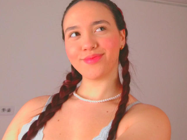 Milliee, con cabello rojo trenzado, piel clara, aro en la nariz, lápiz labial rosa, camiseta azul y collar de perlas, sonriendo ligeramente, mostrando sus pechos, en una foto brillante..