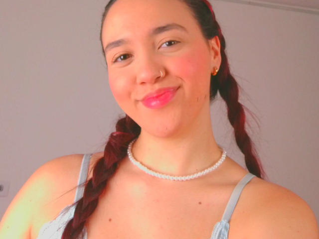 Milliee, con trenzas rojas, lápiz labial rosa, piercing en la nariz, collar de perlas y vestido azul, sonriendo a la cámara, mostrando sus senos. Su piel es clara y es linda..