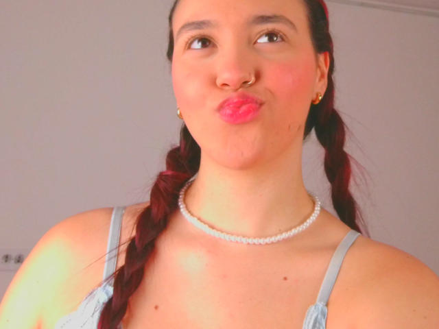 Milliee, con labios rosados y fruncidos, aro en la nariz, cabello rojo trenzado, aretes de oro, collar de perlas y una camiseta gris, mostrando su lindo trasero..