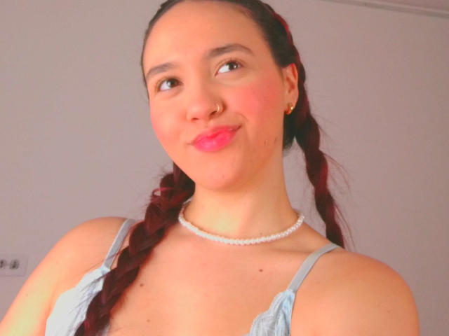 Milliee tem pele clara, cabelo trançado ruivo, um pequeno piercing no nariz, batom rosa, uma blusa azul clara, um colar de pérolas, está sorrindo levemente, e seus seios pequenos estão visíveis..