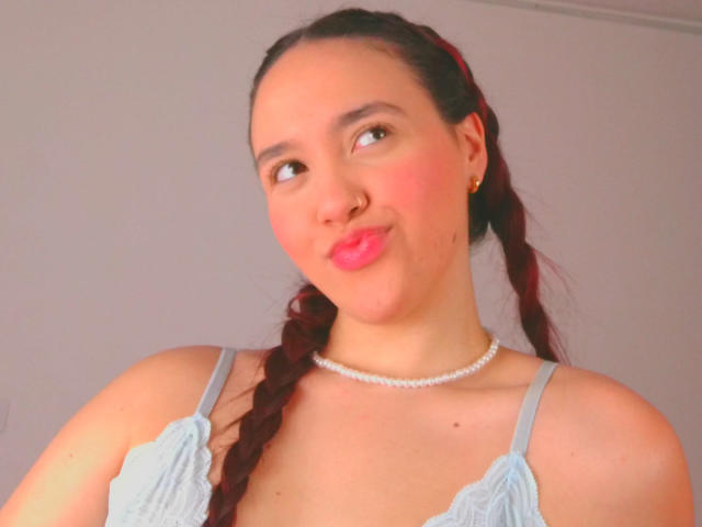 Milliee ha la pelle chiara, rossetto rosa e trecce rosse, indossa una camicetta di pizzo azzurra e una collana di perle, con un piccolo piercing al naso, e sorride leggermente..