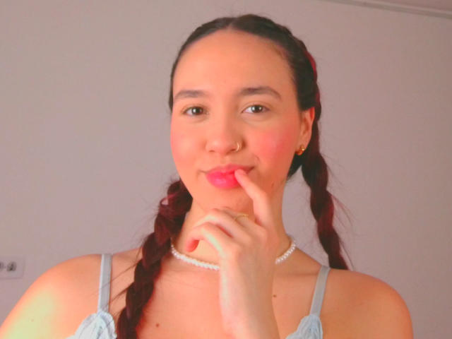 Milliee tem cabelo ruivo trançado, pele clara e está mordendo o dedo, usando batom rosa. Ela veste uma blusa azul, um colar de pérolas e um piercing no nariz..