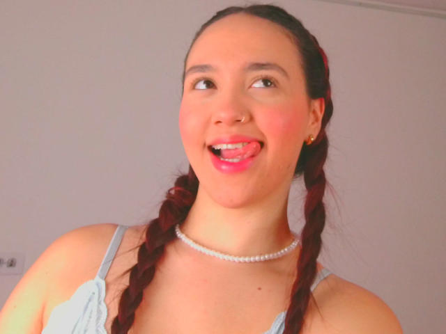 Milliee, con cabello rojo trenzado, un top azul con tirantes finos, un collar de perlas, sacando la lengua, sonriendo, ojos marrones, piel clara, pequeños aretes dorados, en una habitación bien iluminada..