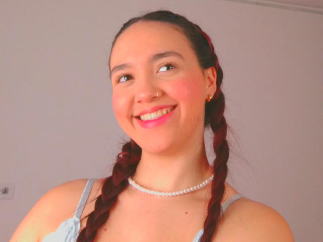 Milliee, com tranças vermelhas, sorrindo, vestindo uma blusa cinza, um colar de pérolas, um piercing no nariz e batom rosa, aparecendo levemente com os seios em um selfie com iluminação suave e quente..