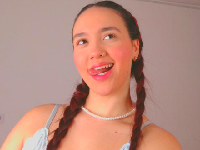 Milliee, com cabelo ruivo trançado, vestido azul claro, colar de pérolas e batom rosa, com a língua de fora de forma divertida, sorrindo para a câmera, olhos castanhos, fofa, atraente..
