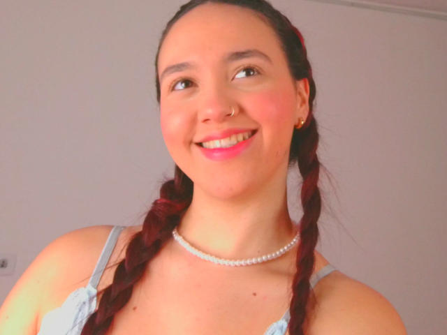 Milliee, com cabelo ruivo trançado, sorrindo, vestindo um vestido azul claro, um colar de pérolas e um piercing no nariz, tem batom rosa e um rosto bonito, mostrando levemente seus seios..