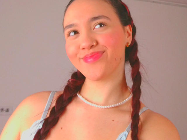 Milliee, avec ses cheveux roux tressés, son rouge à lèvres rose, son anneau de nez, ses boucles d'oreilles dorées, son collier de perles, souriante, portant un haut bleu clair, avec un blush rose pâle sur les joues, mignonne, séduisante, buste..