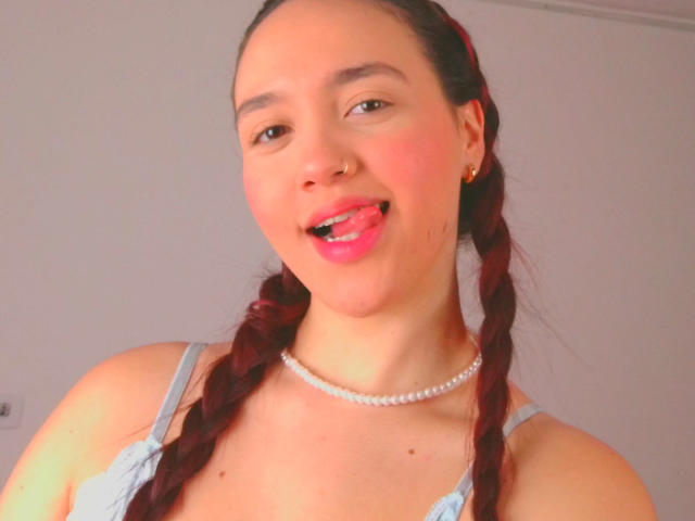 Milliee met gevlochten rood haar, roze lippenstift, tong uitgestoken, parelketting, blauwe jurk, neusring, lachend, lichte huid, kleine oorbellen, bovenlichaam, schattig, sexy..
