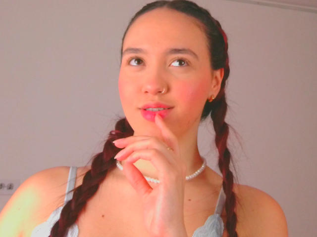 Milliee, com cabelos castanhos trançados, vestindo um vestido azul claro, tocando seus lábios rosados com o dedo, usa um piercing no nariz e um colar de pérolas, e parece adorável..