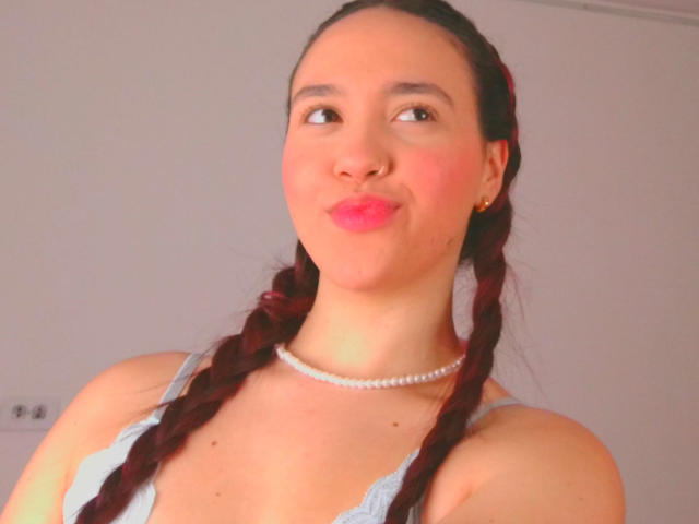 Milliee mit Zöpfen, pinkem Lippenstift und einer Perlenkette, die zur Seite schaut und ein schmollendes Gesicht hat, trägt eine hellblaue Spitzenbluse, die ihre Brüste zeigt..