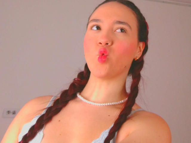 Milliee, avec des couettes, un haut gris clair, un collier de perles et des lèvres roses, regarde sur le côté, exhibant sa poitrine et un joli visage. Éclairage doux et chaleureux..