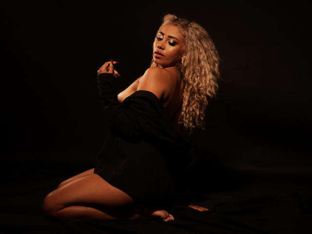 LilithWalker, with blonde curls, sits on black fabric, wearing an off-shoulder black shirt, showing her bare ass and tits, looking over her shoulder with a sultry expression.