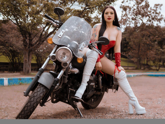 KatalinaJohnson with tattoos, red latex dress, white boots, red gloves, sitting on black motorcycle, outdoors, showing her ass, long black hair, serious expression, motorcycle in front.