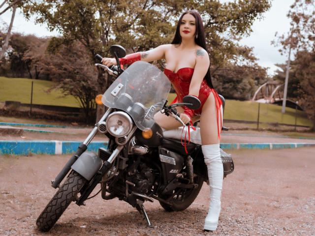 KatalinaJohnson with big boobs, red latex corset, white thigh-high boots, and red gloves, sitting on a black motorcycle, in a park, with a tattoo on her right arm.