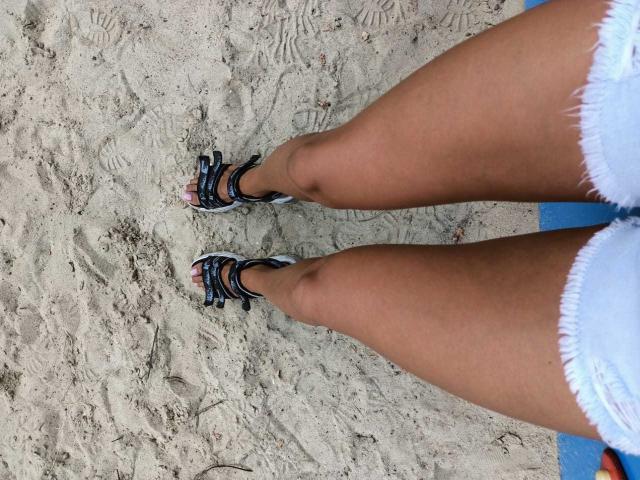 CoupleForFuns' tan thighs in white frayed shorts, wearing black and white strappy sandals, standing on sandy beach.