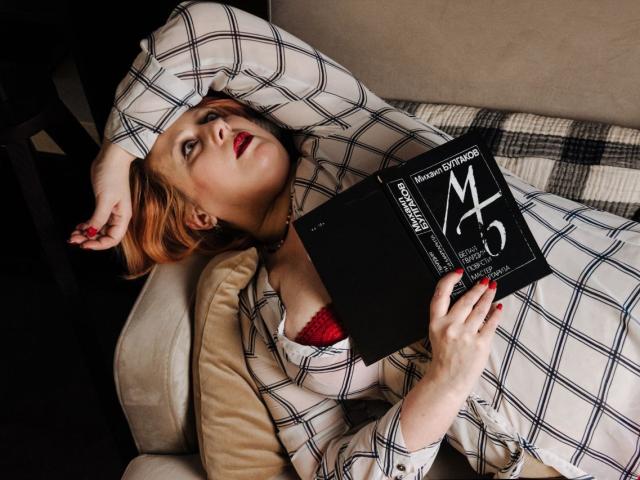 NikolRedMilf con un sujetador de encaje rojo, una camisa de cuadros, acostada en el sofá, leyendo un libro negro, uñas rojas, lápiz labial rojo, cabello rubio, mano en la cabeza, mirando hacia arriba, relajada..
