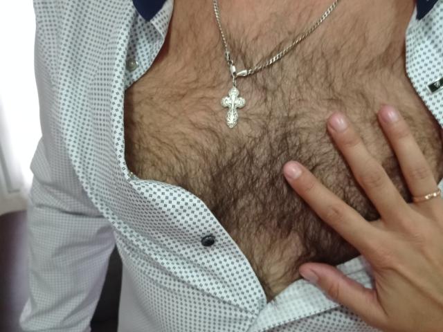 CoupleForFuns's chest with a lot of dark, curly body hair. They're wearing a checkered shirt, a cross necklace, and a ring. CoupleForFuns's hand rests on the chest.